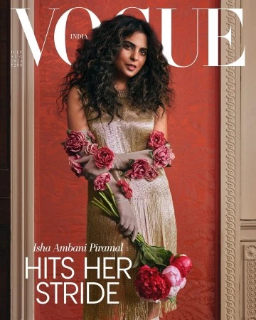 A woman with long dark curly hair wearing a gold dress and sheer gloves adorned with pink roses holds a bouquet of flowers in a magazine cover.
