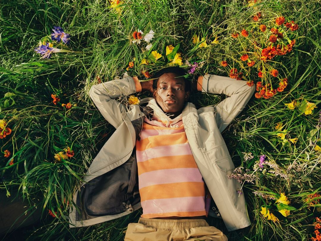 Person lies in the grass surrounded by wildflowers wearing a light-colored jacket and a striped shirt. Relaxing in a field of colorful blossoms.