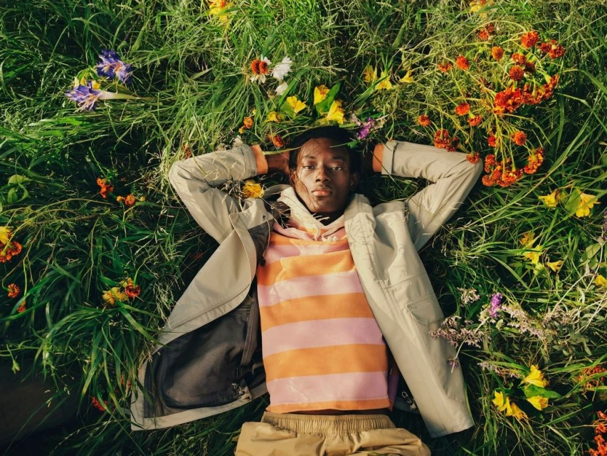 Person lies in the grass surrounded by wildflowers wearing a light-colored jacket and a striped shirt. Relaxing in a field of colorful blossoms.