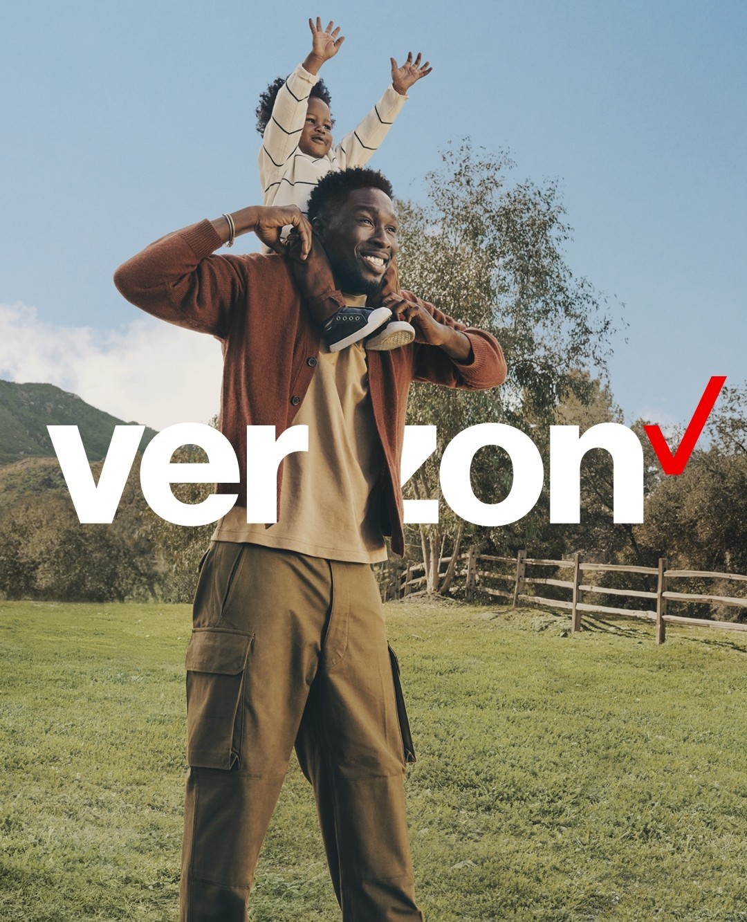 A smiling African-American man carries a young child on his shoulders in a grassy, outdoor setting with the Verizon logo prominently displayed.