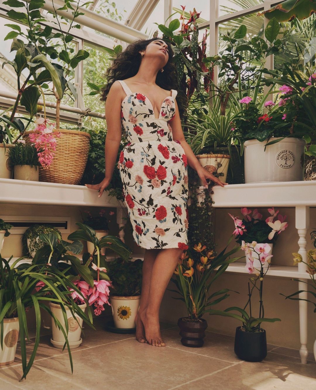 Woman in a floral dress stands barefoot in a sunlit greenhouse filled with various potted plants on shelves and on the floor.