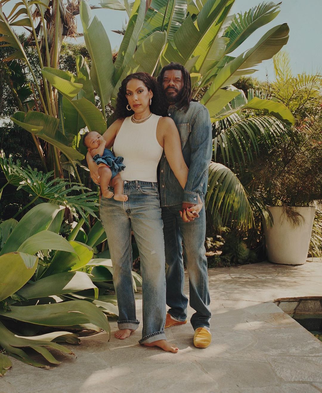 A family portrait featuring a woman holding a baby, standing next to a man, all in denim and light-colored clothes, with lush tropical foliage.
