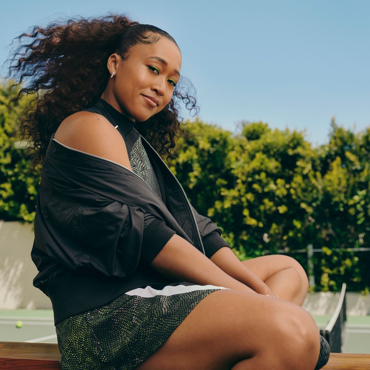 Attractive young woman sits on a bench on a tennis court. She is wearing a black jacket and a patterned dress. Outdoor setting, daytime.