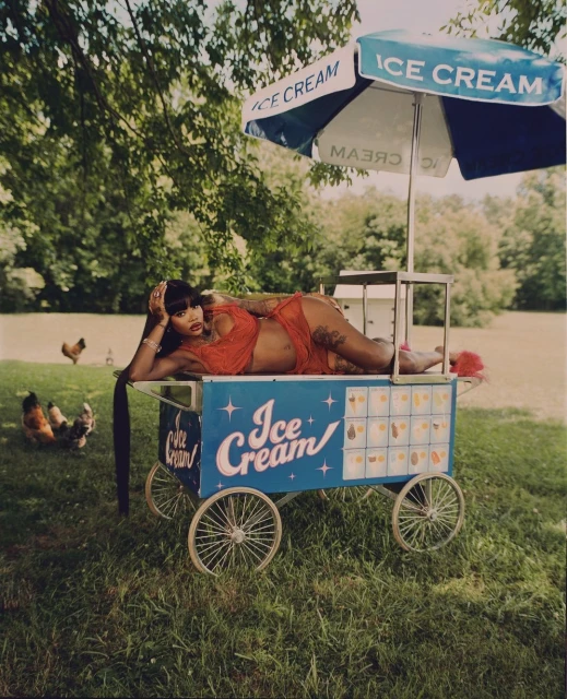 Adult woman in red negligee lounging atop an ice cream cart under an umbrella in a grassy field with scattered chickens.