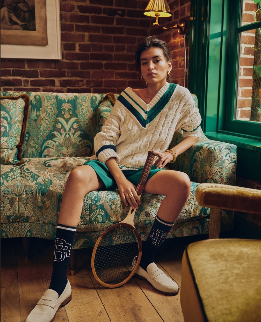 Woman in tennis sweater poses with racket on floral couch in indoor setting with brick wall, window, loafers, and patterned socks.