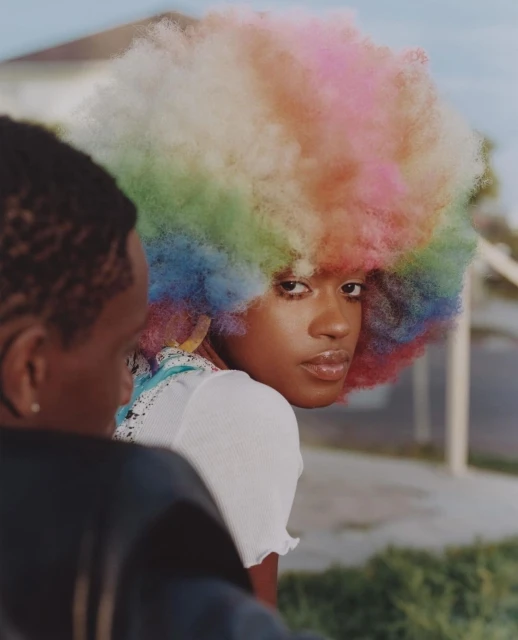 A vibrant photo features a young African American woman with a colorful afro, complemented by a white top, next to another person outdoors.