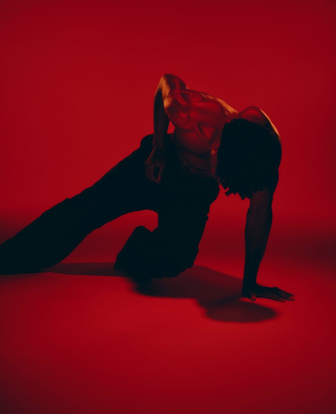 Male dancer in black pants performs a contorted pose in a red studio lit by red lighting, showcasing muscular physique.