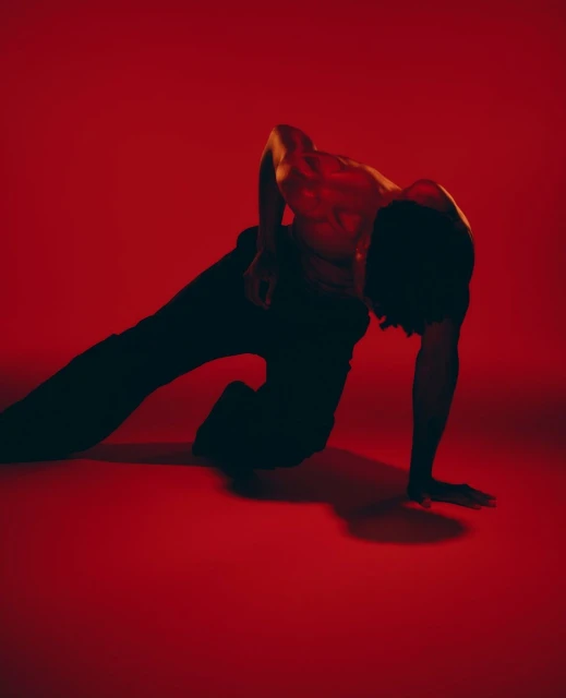 Male dancer in black pants performs a contorted pose in a red studio lit by red lighting, showcasing muscular physique.