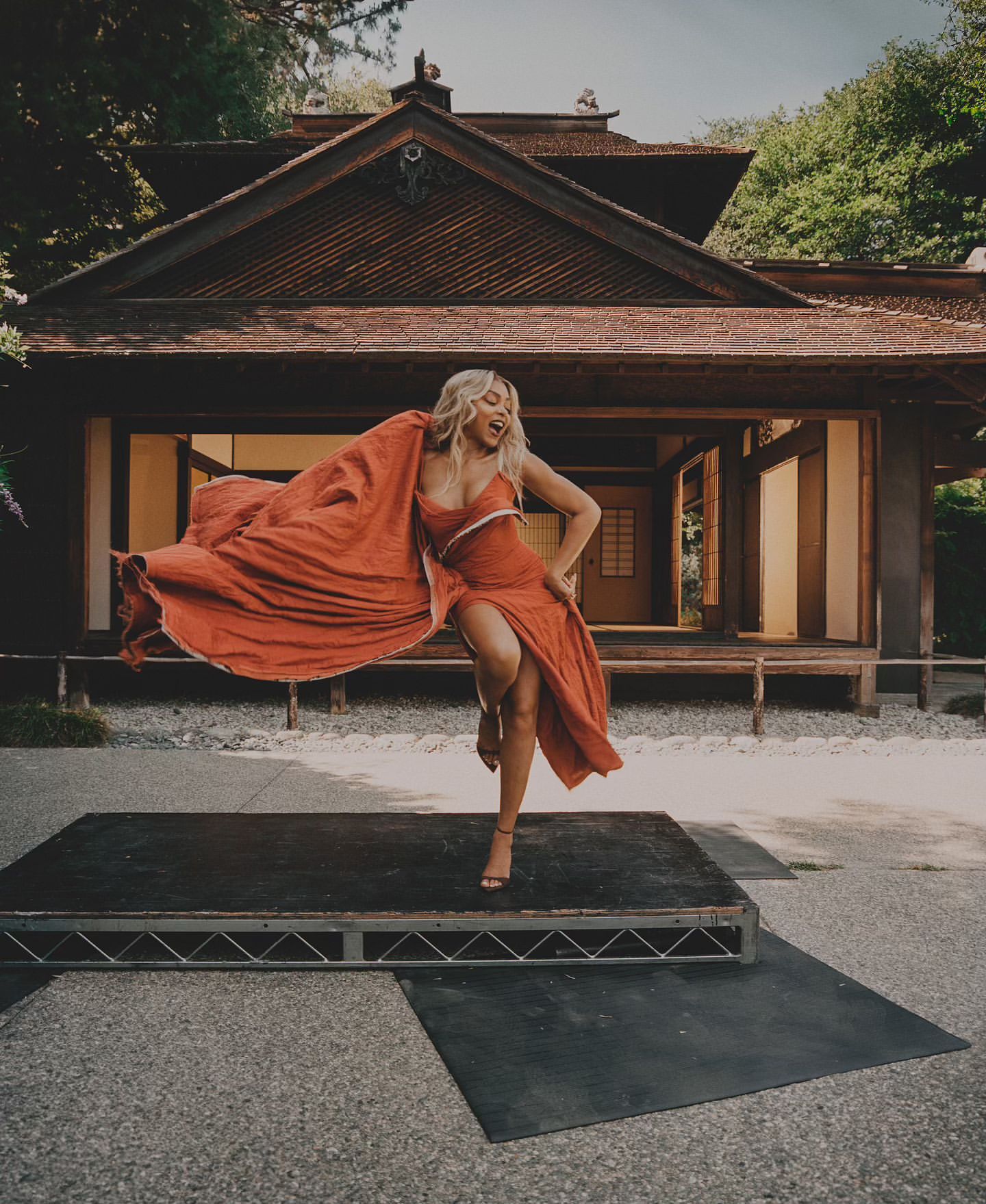 Adult woman with blonde hair wearing a red dress dancing on a black stage in front of Japanese style house, and green plants.