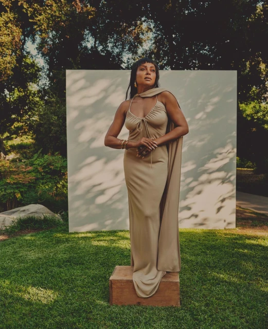 Elegant woman in a long dress posing on a wooden block in a lush green outdoor setting with trees and a light-colored backdrop.
