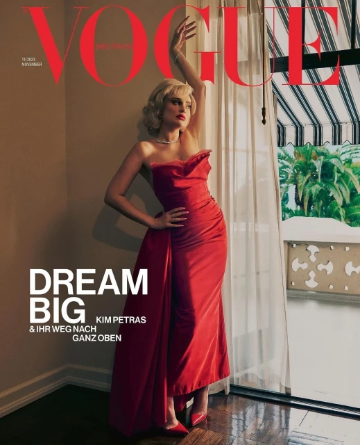 Vogue cover featuring Kim Petras in a red strapless dress, standing near a window with white curtains and a view of palm trees.