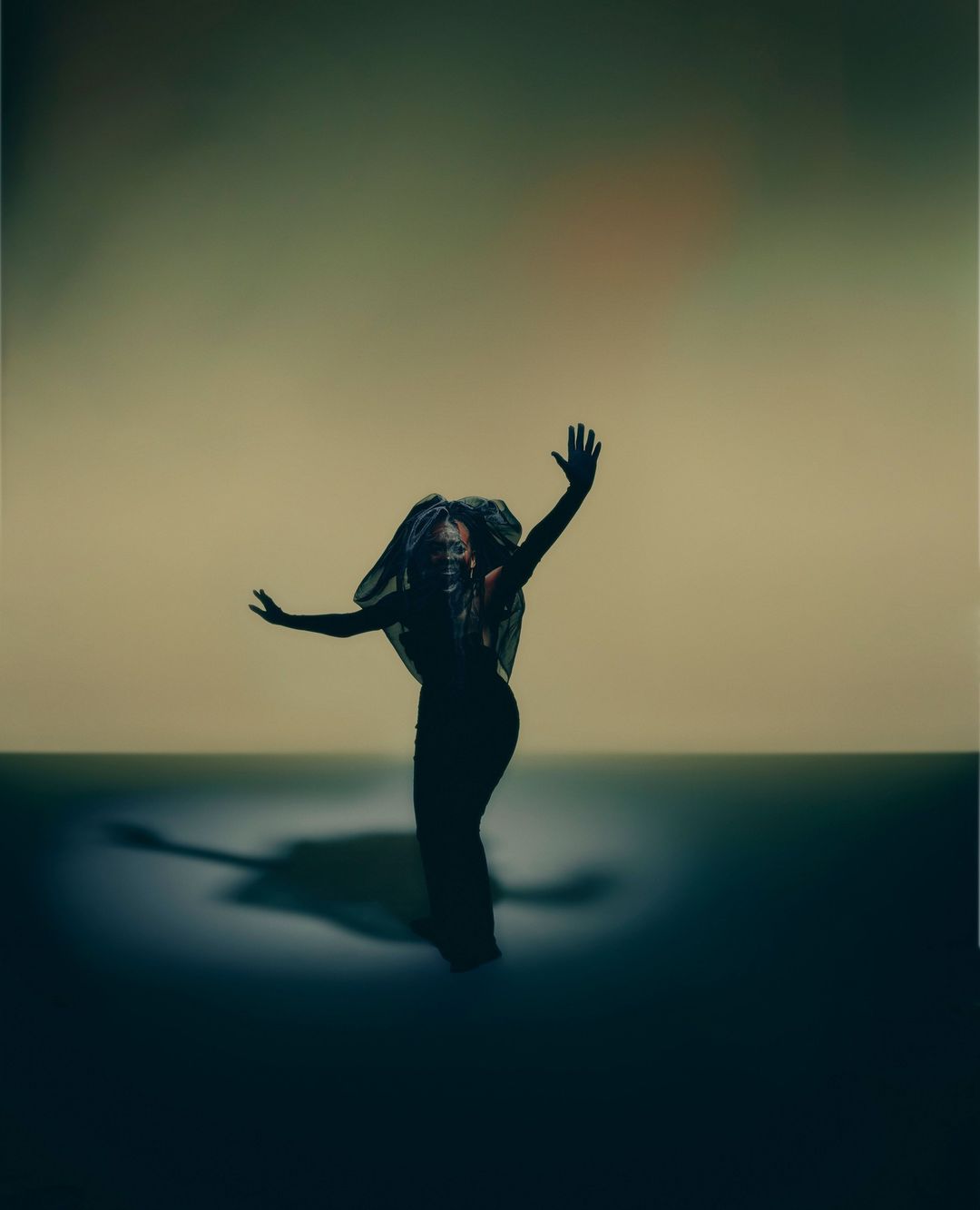 Person in black gown and headdress poses in a photo studio with arms outstretched, dramatically lit against a neutral backdrop.