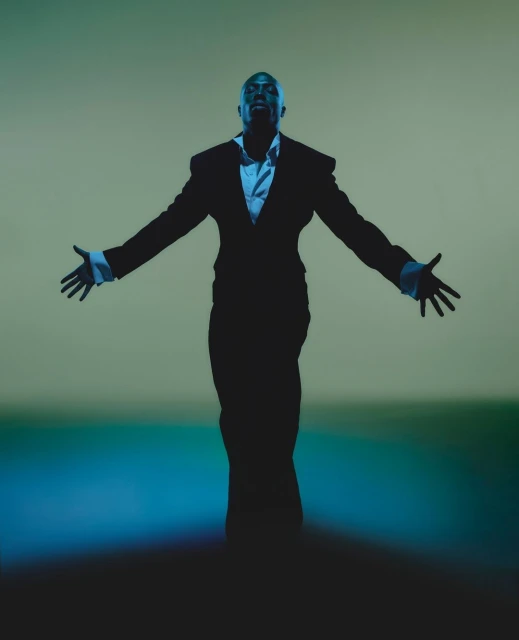 A bald man in a black suit stands with arms outstretched against a green gradient background in a studio lighting environment.