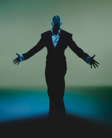 A bald man in a black suit stands with arms outstretched against a green gradient background in a studio lighting environment.