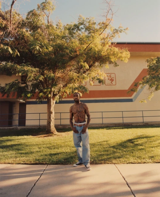 Man with tattoos posing under a large tree in front of a building on a sunny day, wearing jeans and sneakers in an outdoor environment.