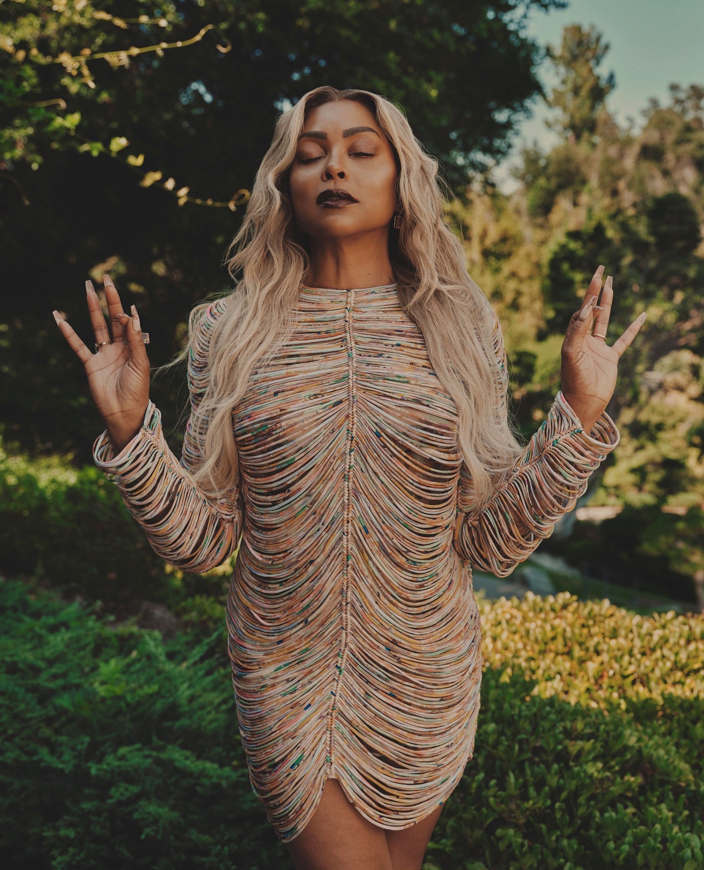 Woman with blonde hair in a beaded mini dress posing with hands in mudra in outdoor green setting. Fashion, style, beauty portrait.