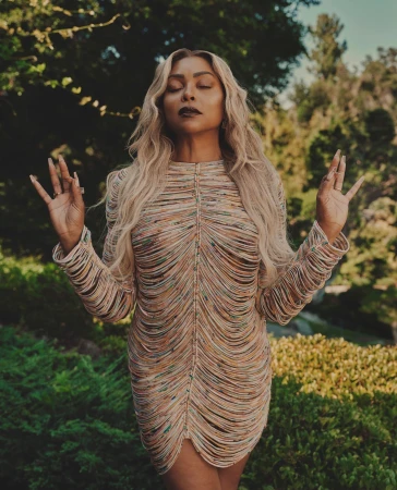 Woman with blonde hair in a beaded mini dress posing with hands in mudra in outdoor green setting. Fashion, style, beauty portrait.