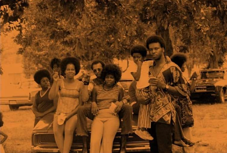 Outdoor shot of a group of African American people, including men, women, and children, gathered near a vintage car with trees behind them.