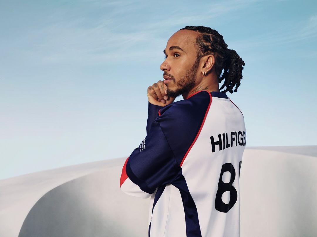 Lewis Hamilton in a Tommy Hilfiger jersey, looking left in a desert environment with sand dunes under a pale-blue sky.