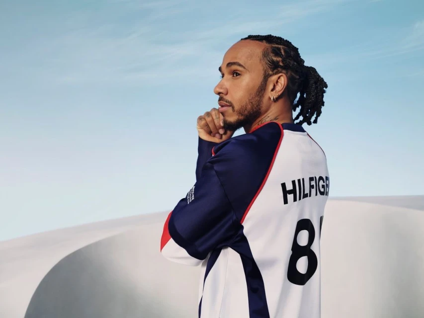 Lewis Hamilton in a Tommy Hilfiger jersey, looking left in a desert environment with sand dunes under a pale-blue sky.