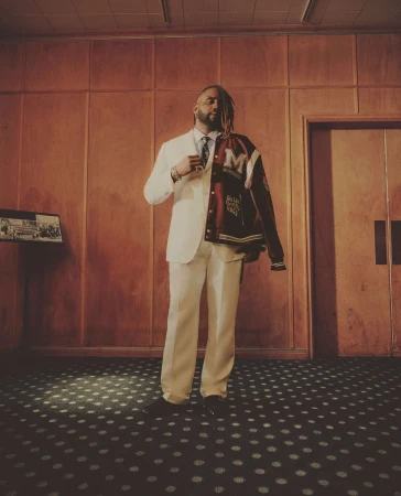 Black man in a white suit with a burgundy varsity jacket draped over his shoulder, photographed by Micaiah Carter.