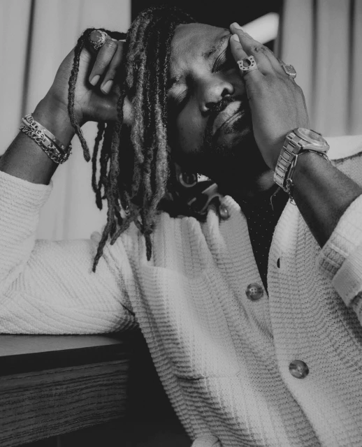 Black and white portrait of a man with dreadlocks wearing a textured white shirt and rings, with hands on head, eyes closed.