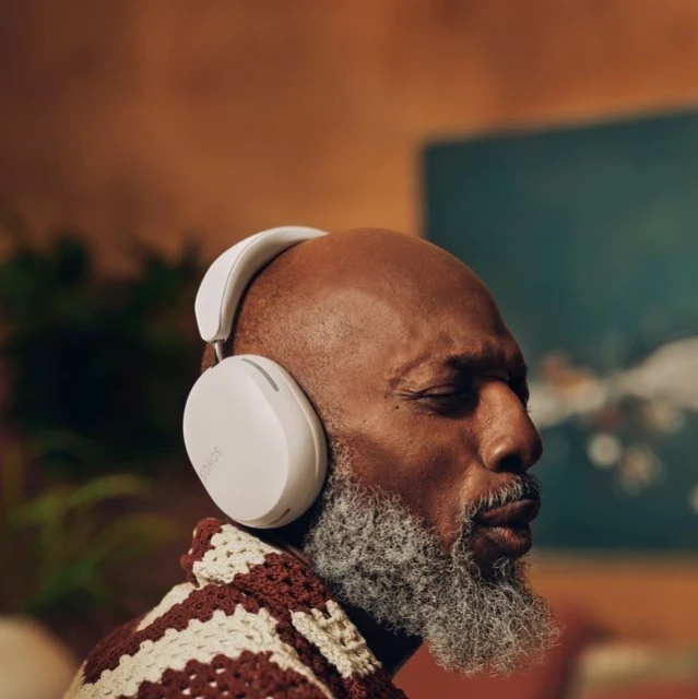 Mature African-American man with closed eyes wearing white headphones and a brown crocheted sweater in a softly lit indoor setting.