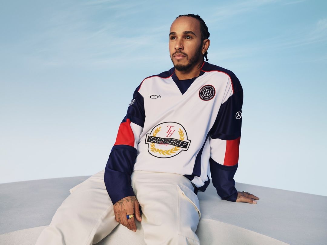 Lewis Hamilton poses in a Tommy Hilfiger shirt and white pants against a blue sky, seated on a white block in a medium shot.