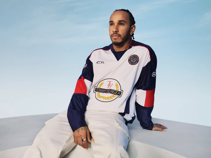 Lewis Hamilton poses in a Tommy Hilfiger shirt and white pants against a blue sky, seated on a white block in a medium shot.