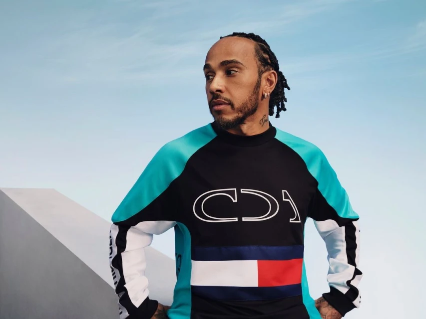 Lewis Hamilton poses in a teal and black top featuring the Tommy Hilfiger logo against a gradient light blue sky backdrop.