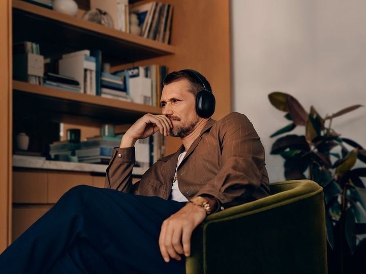 Man wearing headphones and brown shirt seated in an armchair in a room with a bookshelf and houseplant