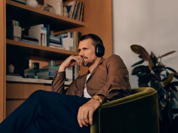 Man wearing headphones and brown shirt seated in an armchair in a room with a bookshelf and houseplant