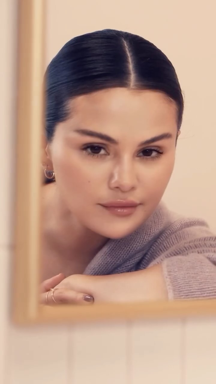Selena Gomez looking into a mirror, leaning on folded arms with styled dark hair and soft makeup, photographed by Micaiah Carter.