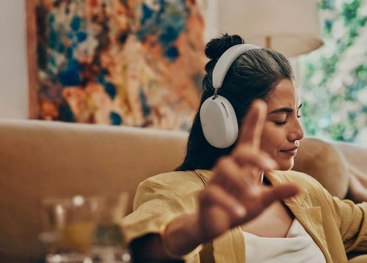 Adult woman listens to white headphones indoors, relaxing on the sofa in a yellow shirt with abstract art in the background.
