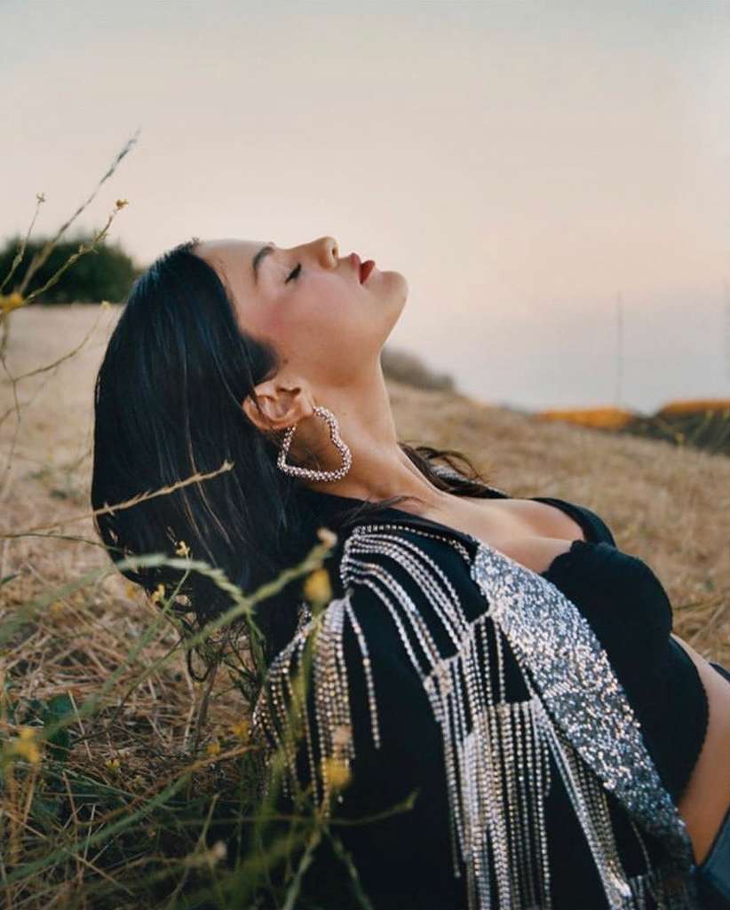 Woman in black top and silver fringed jacket poses in grassy field with head tilted back and heart-shaped earrings, lit by soft sunlight.