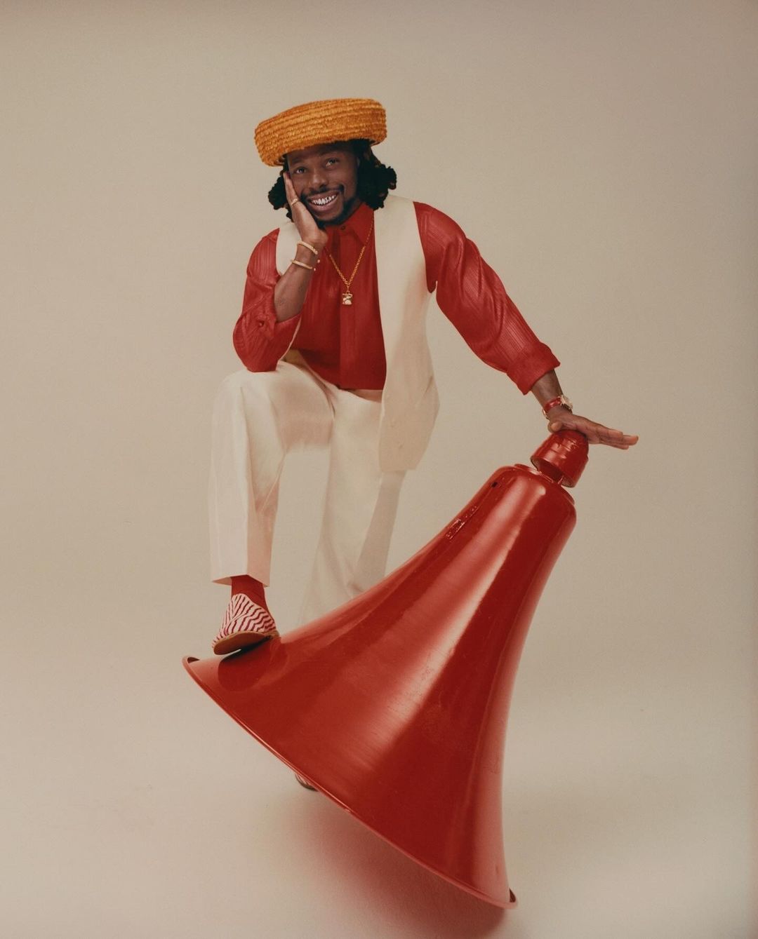 Adult male model wearing a hat, red shirt, and white pants poses with a red object on a neutral background in a studio shot.
