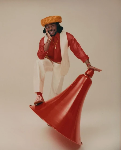Adult male model wearing a hat, red shirt, and white pants poses with a red object on a neutral background in a studio shot.