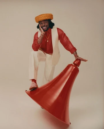 Adult male model wearing a hat, red shirt, and white pants poses with a red object on a neutral background in a studio shot.