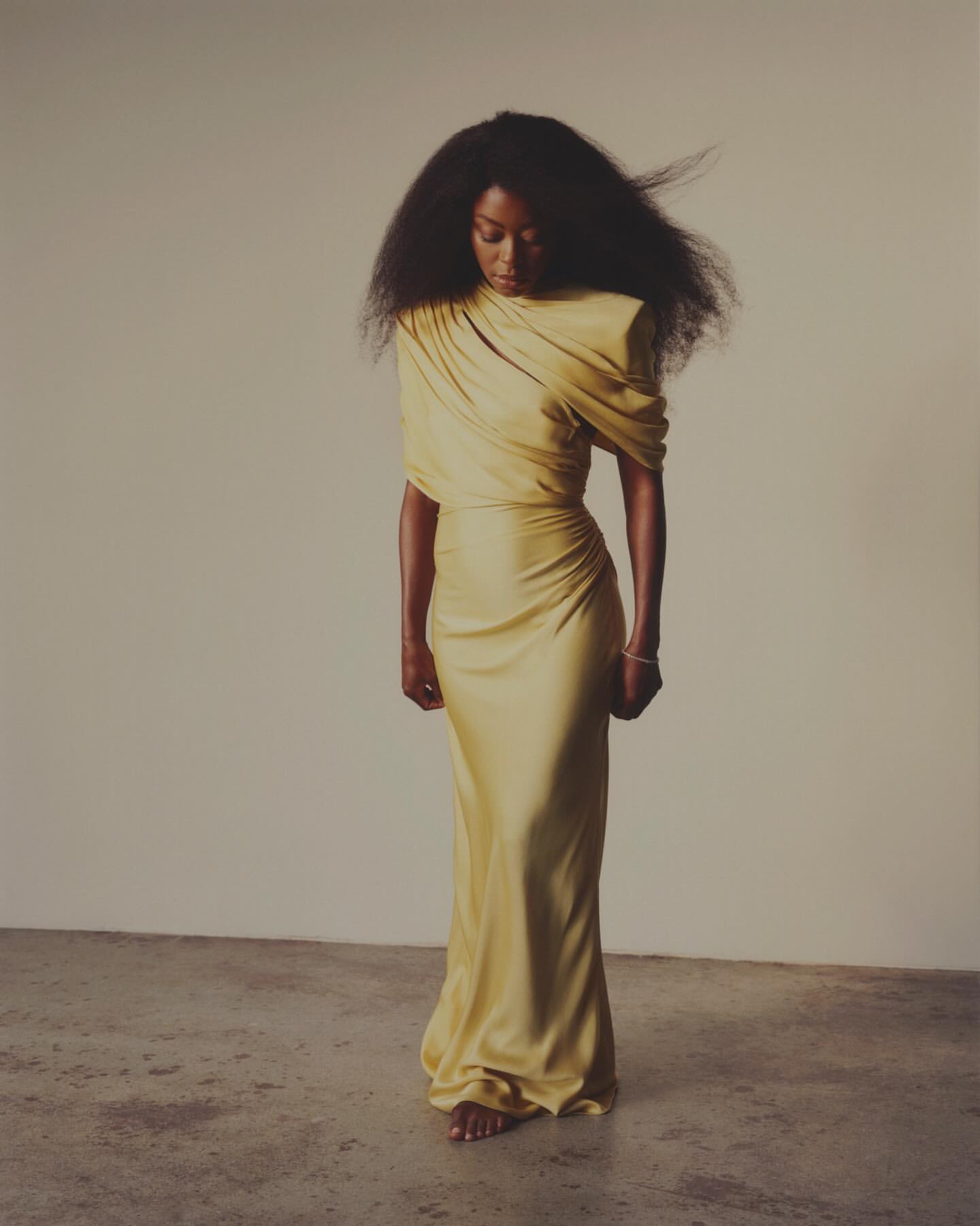 Elegant African-American woman in a draped yellow gown posing in a studio with neutral background, highlighting fashion and beauty.