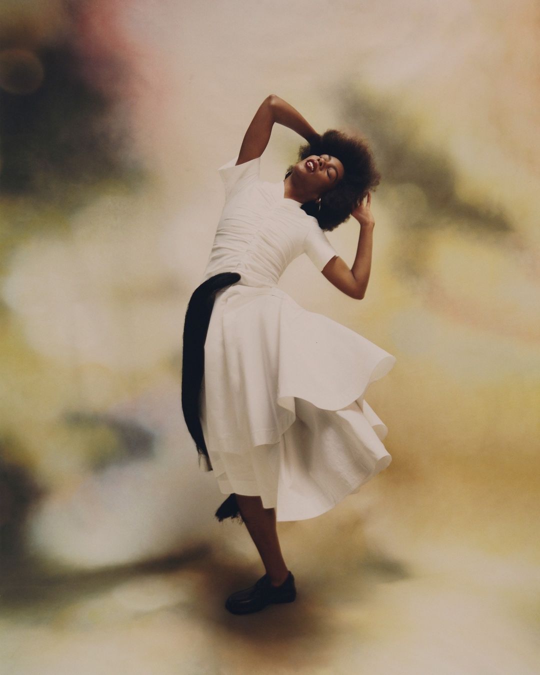 Elegant woman in a white dress poses with afro hair and a black sash against a blurred studio backdrop, showcasing movement and grace.