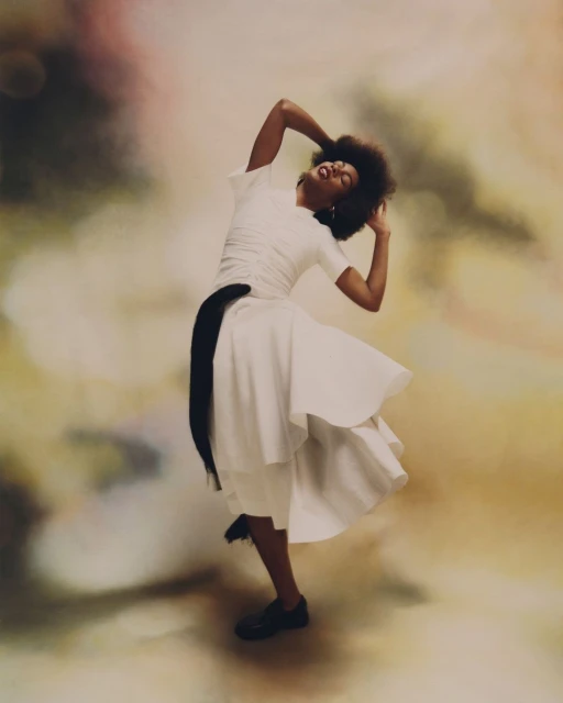 Elegant woman in a white dress poses with afro hair and a black sash against a blurred studio backdrop, showcasing movement and grace.