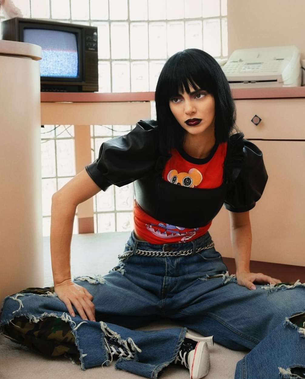 An adult female model with dark hair and makeup poses sitting on the floor in ripped denim jeans, in an indoor studio with a vintage TV.