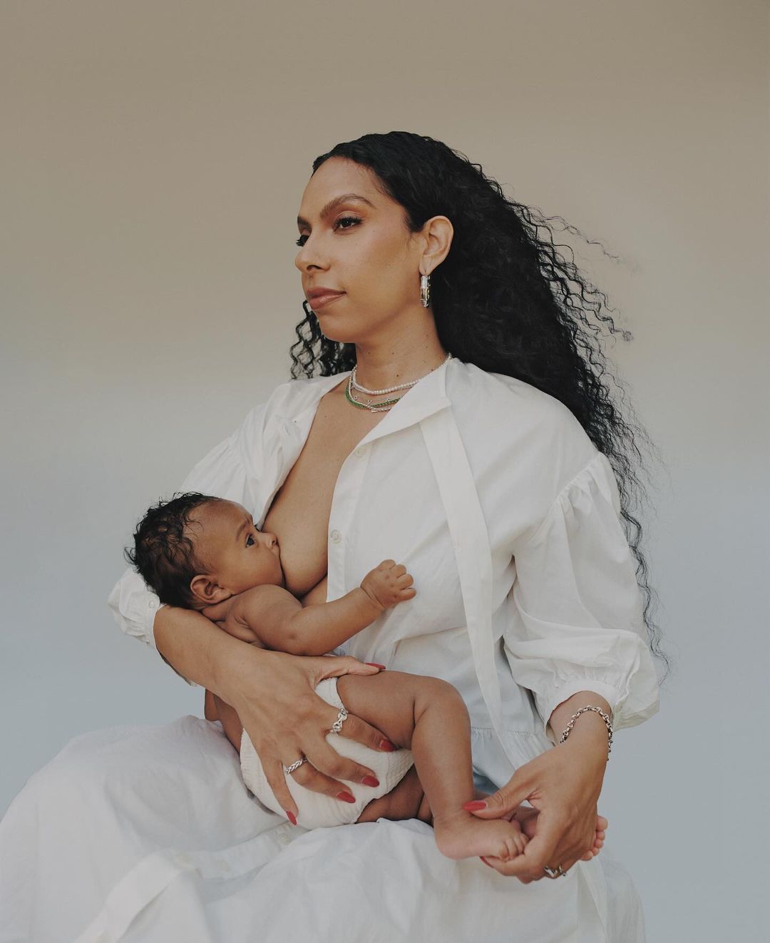 Woman breastfeeding her baby in a studio, wearing a white blouse and jewelry, neutral background, natural light, warm color grading.
