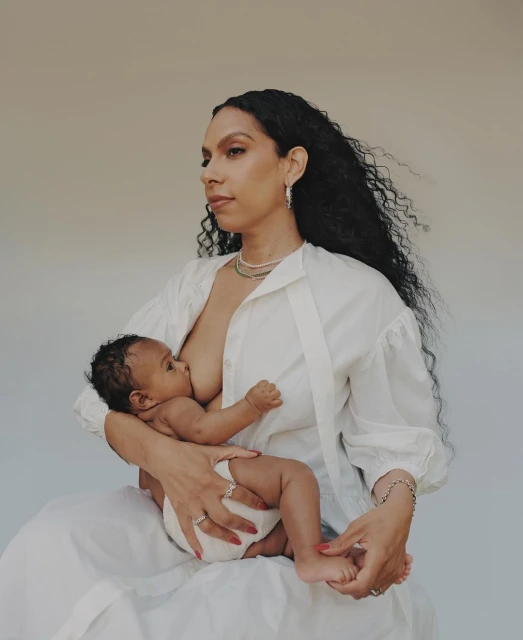 Woman breastfeeding her baby in a studio, wearing a white blouse and jewelry, neutral background, natural light, warm color grading.