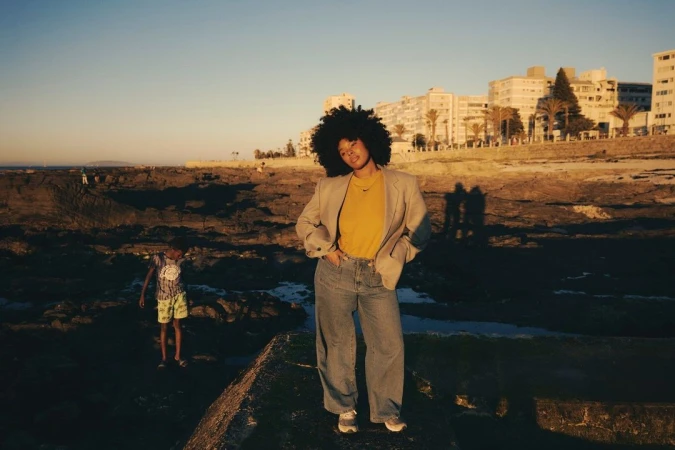 Black woman with voluminous afro hair wearing an oversized blazer, yellow top, and blue jeans standing outdoors near a rocky shore.