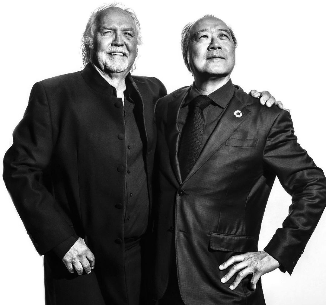 Black-and-white portrait of two adult men in dark suits, standing side by side, looking up, against a bright white backdrop.