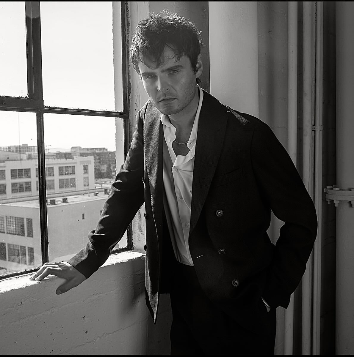 Monochrome portrait of a man in a suit looking at the camera next to a window with buildings in the background. 