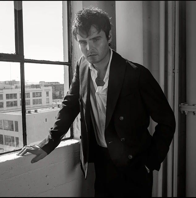 Monochrome portrait of a man in a suit looking at the camera next to a window with buildings in the background. 