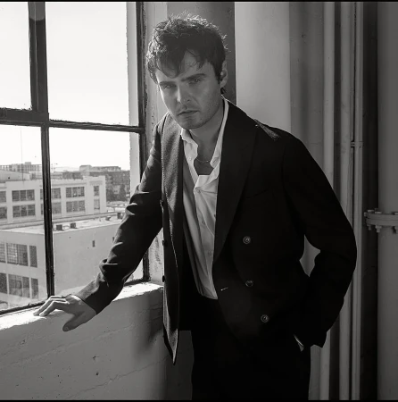 Monochrome portrait of a man in a suit looking at the camera next to a window with buildings in the background. 
