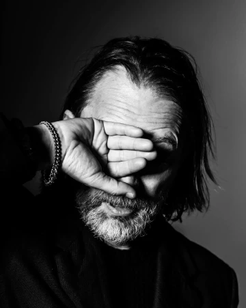 Close-up monochrome portrait of a man with dark hair and beard covering his eyes with his hand.